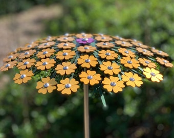 May include: A close-up of a golden metal flower sculpture with a purple center. The sculpture is made up of many small flowers arranged in a circular pattern.