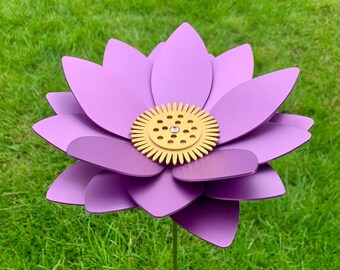May include: A large, purple metal flower garden stake with a yellow center and a small, clear gem. The flower has multiple layers of petals and is set against a backdrop of green grass.