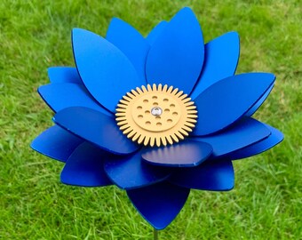 May include: A blue metal flower with a gold gear in the center. The flower is on a green grassy background.