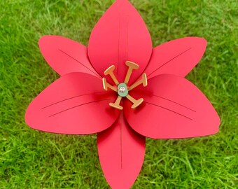 May include: A red paper flower with a gold center. The flower has five petals and is sitting on a green grassy surface.