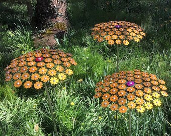 May include: Three metal sculptures of flowers with orange and yellow petals and a purple center. The sculptures are standing in a grassy field.