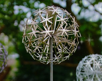 May include: A silver metal sculpture of a dandelion with a starburst design. The sculpture is on a tall metal stem and is surrounded by a blurred green background.