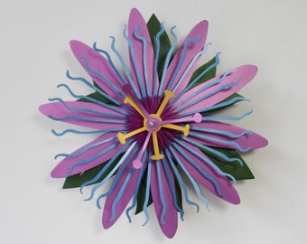 May include: A wall clock shaped like a passion flower with layered purple petals, wavy light blue filaments, and dark green leaves. The center features a yellow and purple structure with clock hands, including a yellow hour hand and a purple minute hand.