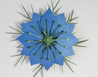 May include: A layered paper craft flower featuring vibrant blue petals arranged in a circular pattern. The center displays a green star-shaped structure with thin, radiating stamens tipped with small green dots, surrounded by thin green foliage.