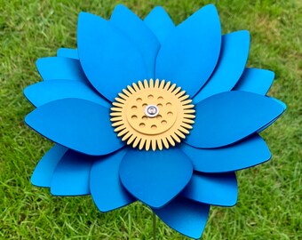 May include: A large, blue metal flower garden stake with a yellow center. The flower has multiple layers of petals and is set against a backdrop of green grass. The flower is a decorative garden accent.