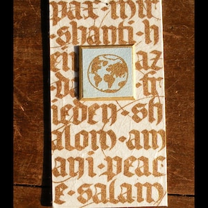 May include: A white card with gold lettering that reads "pax-wir shanti-h el az ieden-sh alow-am ayi-peac e-salam". A small square frame with a blue background and a gold globe inside is centered on the card.
