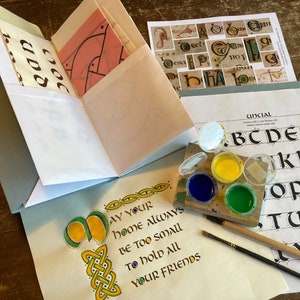 May include: A calligraphy practice set featuring a grey folder with instructional pages, a sheet with Uncial alphabet letters, and a sheet with the text "May your home always be too small to hold all your friends." Includes a small paint palette and brushes.