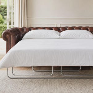 May include: A brown leather sofa bed featuring a tufted backrest with rolled arms and nailhead trim. The sofa is extended into a bed position with a white mattress, two white pillows, and a visible silver metal frame base underneath.