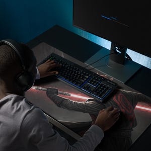 May include: A large rectangular desk mat featuring a printed graphic of a character holding a glowing red double-bladed lightsaber. The mat is positioned under a black computer keyboard with blue backlighting and a computer mouse.
