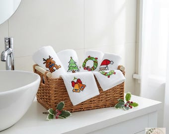 以下が含まれることがあります： 茶色の編み込み籐かごの中に、トナカイ、クリスマスツリー、リース、サンタクロース、金色の鐘などのカラフルなクリスマスのモチーフが刺繍された白いハンドタオルが数枚入っており、ヒイラギの小枝が添えられています。