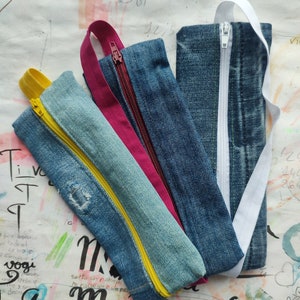 以下が含まれることがあります： ジッパーと布製のストラップが付いたデニム製のペンケースが3つ。ケースは、黄色のジッパー付きケースが下、ピンクのジッパー付きケースが中央、白いジッパー付きケースが上に並んでいます。