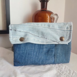 May include: A small blue denim pouch with a flap closure and two metal poppers. The flap is made of a lighter shade of denim and has a frayed edge.