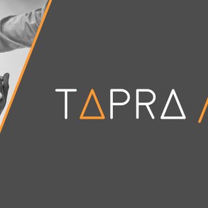 May include: A black and white image of a group of people holding hands in a circle. The image is cropped to show only the hands and arms of the people. The background is a dark gray color. The text "TAPRA / management & engineering" is in white and orange on the right side of the image.