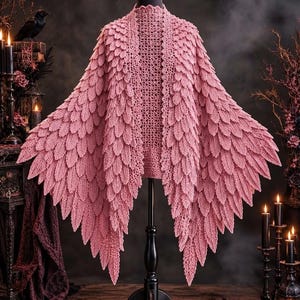 Peut inclure: Un châle en crochet rose poudré présentant une texture dense et superposée rappelant des plumes sur tout le vêtement. La pièce est construite avec un motif en maille ajourée au centre du dos et présente de longues franges pointues.