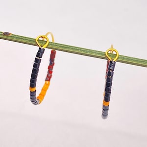 Puede incluir: Un par de pendientes de aro con pequeñas cuentas cilíndricas en negro, naranja y rojo moteado. Cada aro está sujeto a un fino bucle de metal dorado, creando un diseño circular con un patrón repetitivo de cuentas de colores.
