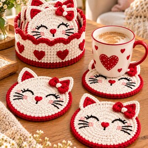 May include: A set of round, cream-coloured crochet cat coasters with red ears, red bows, and embroidered black whiskers. The set includes a matching circular crochet basket with red heart motifs and a white ceramic mug with red heart patterns.
