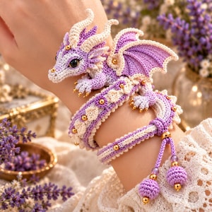 May include: A crochet bracelet featuring a small, lavender and cream-colored dragon with large dark eyes, intricate wings, and gold-toned beads. The band is crafted from braided yarn in matching colors, finished with two dangling, spherical crochet charms.
