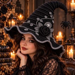 May include: A black crocheted witch hat featuring a wide, wavy brim with a light gray edge. The hat is decorated with a large black crocheted rose, gray leaf accents, and a small white cross emblem. A crocheted spider hangs from the pointed tip.