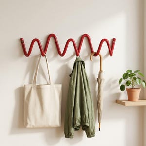 May include: A red, wall-mounted coat rack with a continuous, wavy, serpentine design. The rack features multiple hooks holding a beige canvas tote bag, an olive green jacket, and a closed beige umbrella with a curved wooden handle.