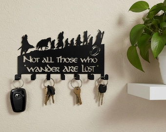 Könnte beinhalten: Ein schwarzer Wand-Schlüsselhalter aus Metall mit einer Silhouette wandernder Figuren, einem Ring-Symbol und dem Text "Not all those who wander are lost". Die Leiste hat sechs Haken, an denen drei verschiedene Schlüsselbunde hängen.