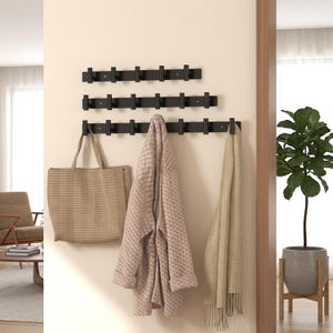 Puede incluir: Tres percheros de pared horizontales de color negro con múltiples ganchos cilíndricos. Los soportes están dispuestos en forma escalonada y sostienen un bolso beige, un cárdigan beige texturizado y una bufanda larga beige.
