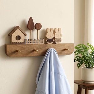 May include: A wooden wall-mounted coat rack featuring three brass hooks and a decorative top shelf. The shelf displays a small wooden house, two stylized trees, a picket fence, and two rabbit figures with light and dark wood grain details.