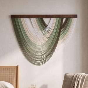 May include: A wall hanging featuring multiple strands of yarn in shades of sage green, cream, and dark brown, suspended from a horizontal dark wood bar. The yarn is arranged in overlapping, draped semicircular loops.