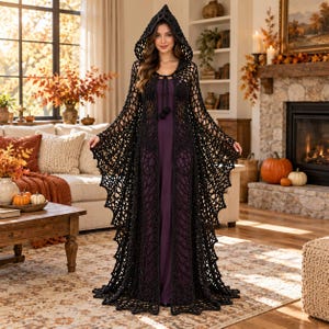 May include: A floor-length black crochet hooded cloak featuring an open-knit spiderweb pattern and a tie closure at the neck, worn over a solid dark purple sleeveless dress. The garment has wide, flowing sleeves and a pointed hood.