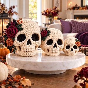 May include: Three cream-colored crocheted skulls of varying sizes arranged on a white marble stand. Each skull features dark eye sockets and is decorated with crocheted flowers and leaves in shades of deep red, orange, and dark green.