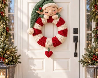 May include: A crocheted Christmas wreath featuring a sleeping elf with a long green hat and pointed ears resting on a red and white striped ring. A small red heart hangs from the bottom of the wreath, which is displayed on a white door.