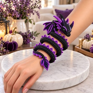 May include: A crochet dragon bracelet featuring a coiled black and purple body with detailed wings, spikes along the spine, and a pointed tail. The creature has a textured yarn construction with a defined head, dark eyes, and small ears.