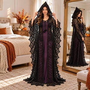 May include: A floor-length black crochet hooded cloak featuring an intricate open-work web pattern, long flared sleeves, and a tie closure at the neck, worn over a solid dark purple sleeveless maxi dress.