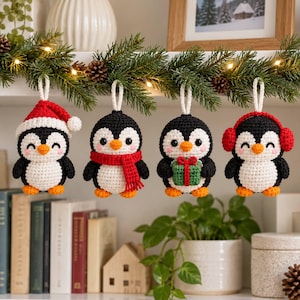May include: Four crocheted penguin ornaments hanging from a green garland. The penguins are black and white with orange beaks and feet. They wear a red Santa hat, a red scarf, hold a green gift box, or wear red earmuffs, each with a white loop.