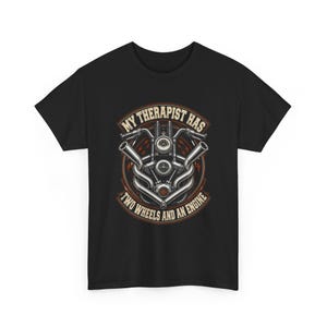 Pode incluir: Uma t-shirt preta de manga curta com um gráfico de um motor de motocicleta e guiador. O texto "MY THERAPIST HAS TWO WHEELS AND AN ENGINE" está disposto num padrão circular em torno da ilustração mecânica em laranja e branco.