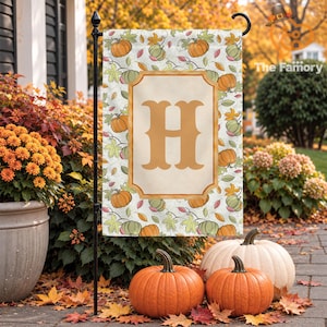 May include: A rectangular garden flag featuring a repeating pattern of orange pumpkins and colourful autumn leaves on a white background. A central tan rectangular frame contains a large, bold, golden-brown letter H in the centre.