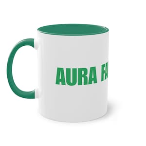 Puede incluir: Una taza de cerámica blanca con interior y asa de color verde. El exterior presenta el texto verde AURA FA impreso en una fuente negrita sin remates. La taza tiene una forma cilíndrica con un acabado liso y un asa curva en el lado izquierdo.