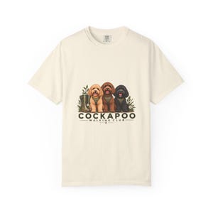 Puede incluir: Una camiseta de manga corta color crema con un gráfico de tres perros Cockapoo con pañuelos verdes. El texto dice COCKAPOO WALKING CLUB EST 2023. Los perros están rodeados de follaje verde y una bota verde sobre un fondo liso.