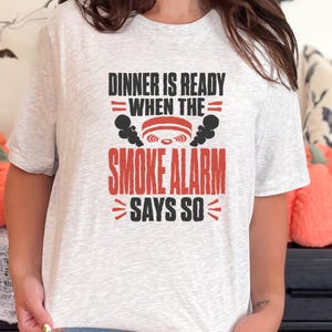 May include: A light gray heathered short-sleeve t-shirt featuring black and red text that reads DINNER IS READY WHEN THE SMOKE ALARM SAYS SO, accompanied by a graphic of a smoke detector with black smoke plumes rising from the sides.