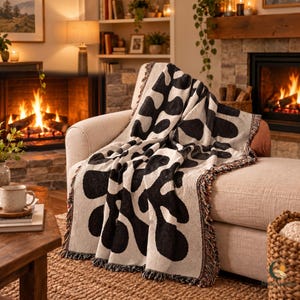 Peut inclure: Un plaid tissé présentant un motif organique abstrait à fort contraste noir et crème. Ce textile rectangulaire possède une surface texturée et est agrémenté de franges décoratives multicolores sur les bords, drapé sur un canapé.
