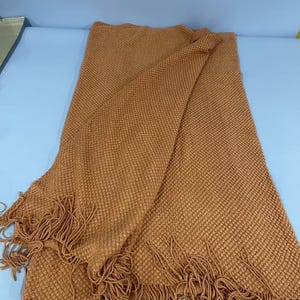 Puede incluir: Una manta tejida de color naranja quemado, de forma rectangular, con un patrón de punto texturizado similar a un gofre en toda la superficie. El borde inferior está rematado con largos flecos a juego a lo largo de todo el ancho.