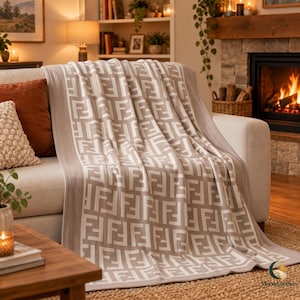May include: A soft, knitted throw blanket featuring a repeating geometric pattern of the letter F in white against a light brown background. The blanket is draped over a light-colored sofa, showcasing its textured fabric and consistent design.