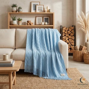 May include: A light blue knit throw blanket featuring a diamond pattern and fringe trim along the bottom edge. The blanket is draped over a beige sofa and includes a small rectangular label in the bottom corner with the text MoonAuraSun.