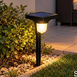 May include: A black solar-powered garden light with a square top panel and a ribbed, translucent glass shade emitting a warm yellow glow. The light is mounted on a cylindrical black post, positioned upright in a garden bed with stones.