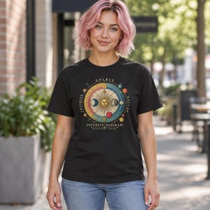 Puede incluir: Una camiseta negra de manga corta con un gráfico circular de un diagrama celestial. El diseño incluye un sol con rostro, fases lunares y esferas planetarias con el texto SPIRIT, NATURE, INFINITE e INFINITE DIAGRAM.