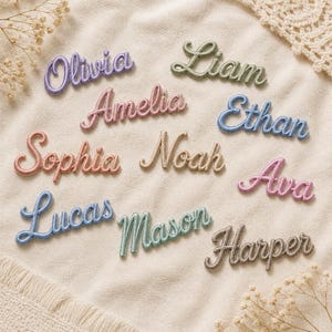 Pode incluir: Uma coleção de emblemas bordados com nomes em várias cores, incluindo roxo, verde, rosa, azul, pêssego, bege e castanho. Cada emblema apresenta um nome em letra cursiva, especificamente Olivia, Liam, Amelia, Ethan, Sophia, Noah, Ava, Lucas, Mason e Harper.