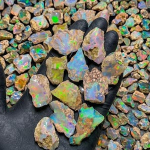 May include: A collection of raw, unpolished opal stones featuring a natural, rough texture and a vibrant play-of-color. The stones display iridescent flashes of green, blue, orange, and pink against a milky, translucent, and earthy base.