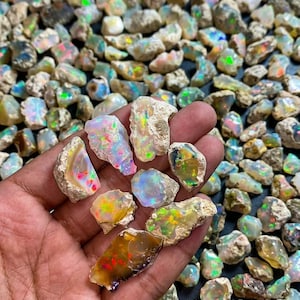 Può includere: Una collezione di pietre di opale grezze e non lucidate caratterizzate da una varietà di colori iridescenti, inclusi bagliori di verde, rosso, arancione e blu. Le pietre hanno forme irregolari e frastagliate con superfici ruvide e traslucide.