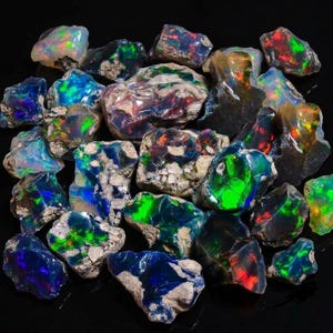 May include: A collection of raw, unpolished opal stones featuring dark, opaque bases with vibrant flashes of iridescent green, blue, and red color play. The stones have irregular, jagged shapes and rough, textured surfaces with visible mineral inclusions.