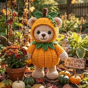 May include: A crocheted teddy bear wearing a pumpkin costume with a hood, green leaves, and a brown stem. The bear has light tan fur, black eyes, and a brown nose. A small wooden sign nearby features the text "AUTUMN PATCH" in black lettering.