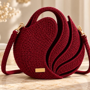 May include: A deep red crochet handbag with a circular silhouette and layered, fan-like side panels. The bag features a matching crochet handle, a detachable shoulder strap with gold-toned metal hardware, and a small rectangular gold-toned metal plaque.